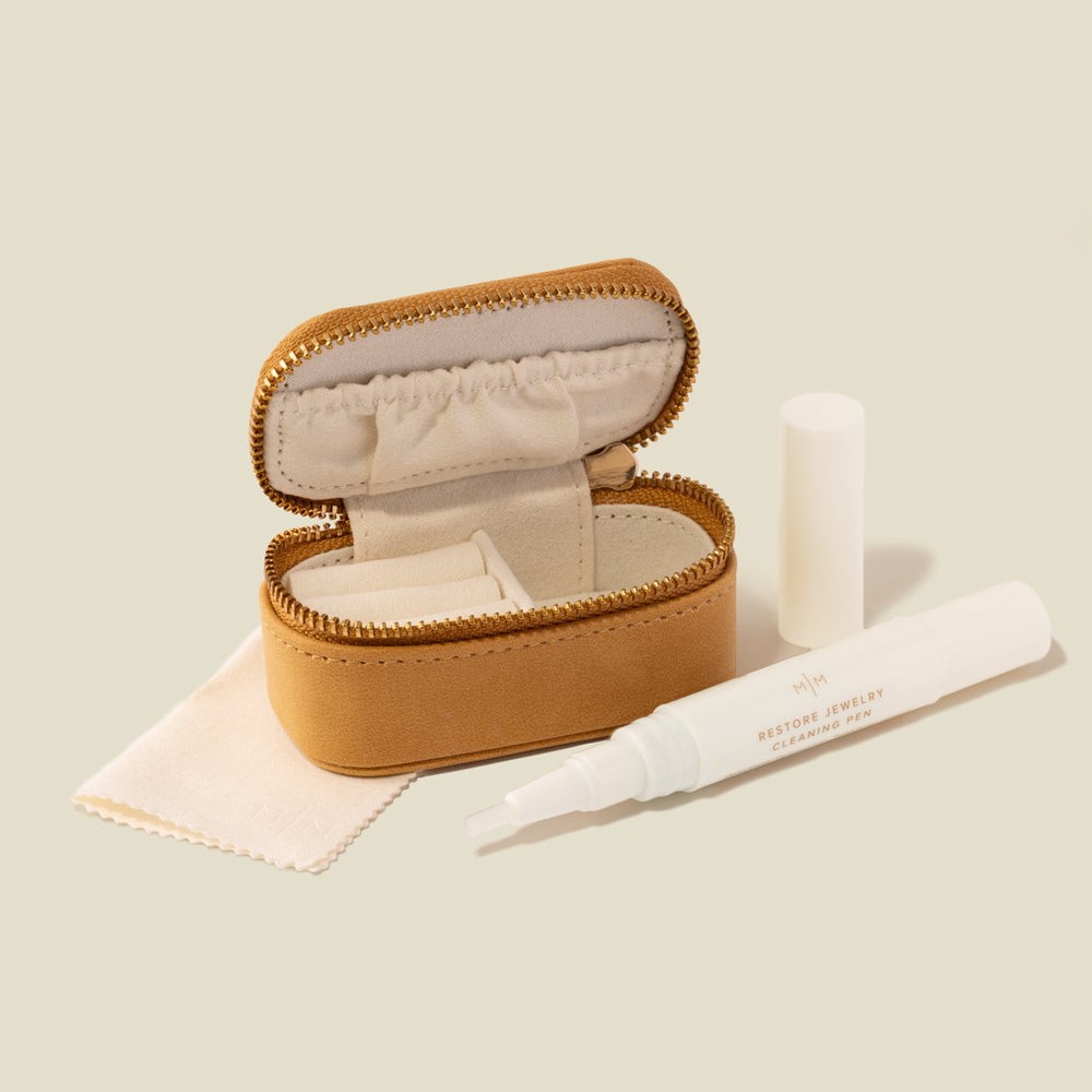 An open tan faux-leather jewelry case with gold-tone zipper, shown with a white polishing cloth and a white jewelry cleaner applicator pen on a soft beige background.