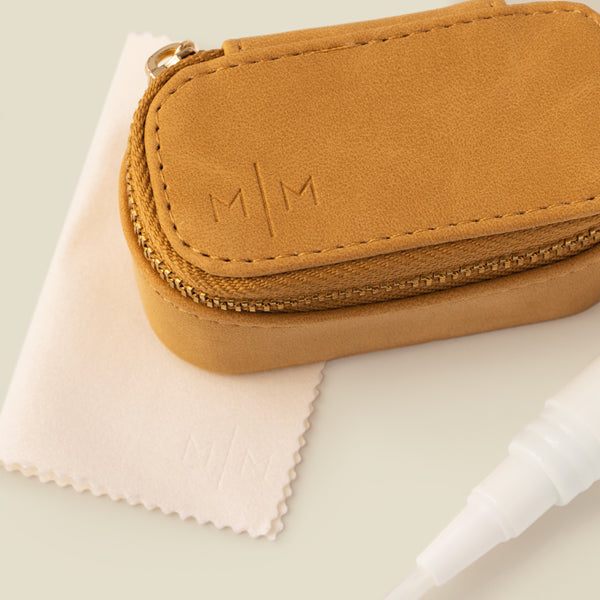 A close-up of a tan faux-leather jewelry case with gold-tone zipper and the logo 'M|M' embossed on top, shown with a white polishing cloth and a white jewelry cleaner applicator pen on a soft beige background.