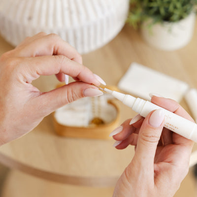 A close-up of a person using a white jewelry cleaner applicator pen on a delicate gold ring, with a table and plant blurred in the background.