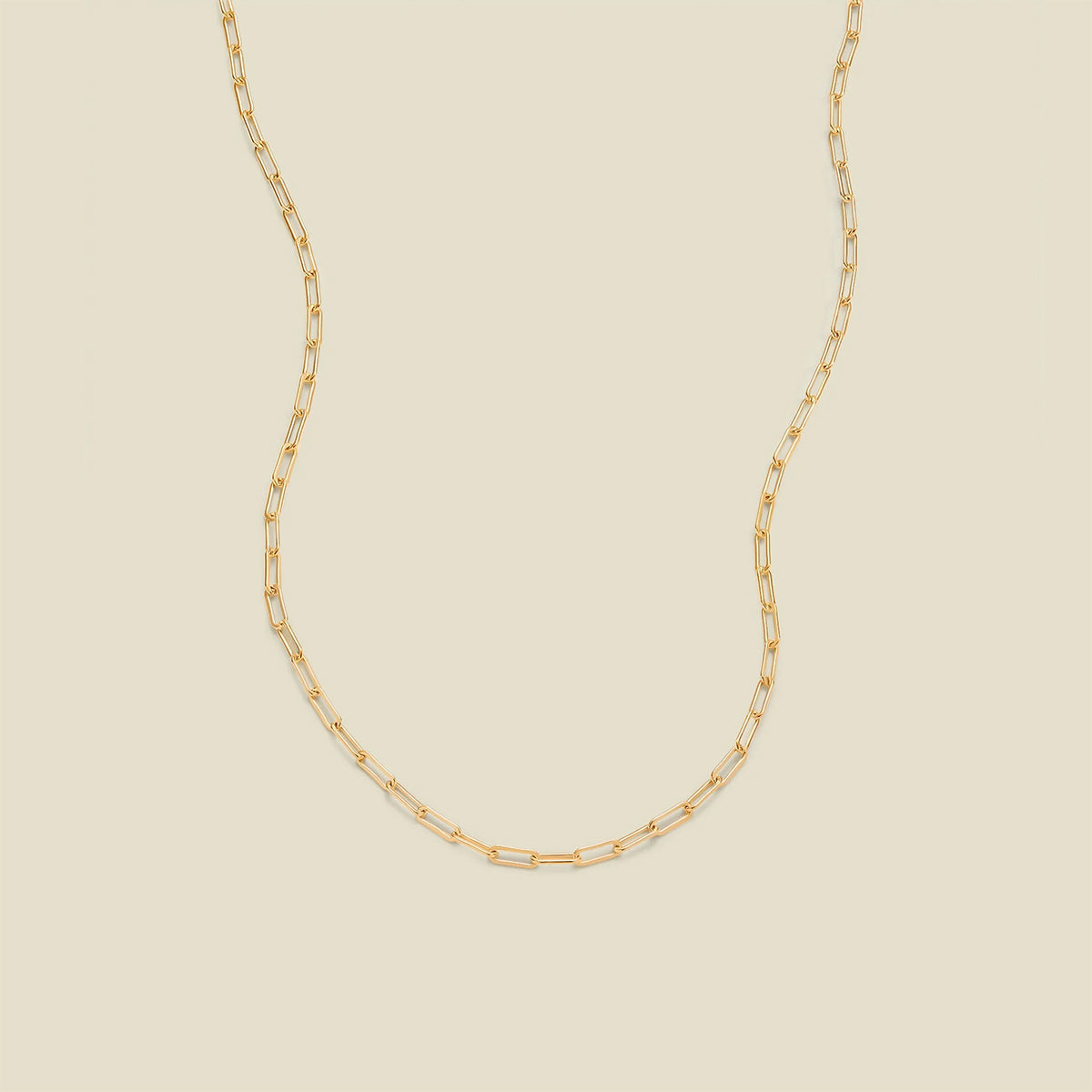 A gold paperclip chain necklace draped in a U-shape against a solid beige background.