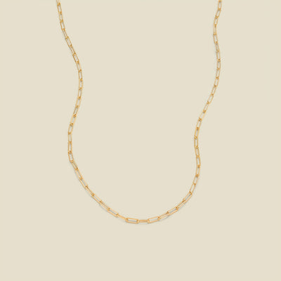 A gold paperclip chain necklace draped in a U-shape against a solid beige background.