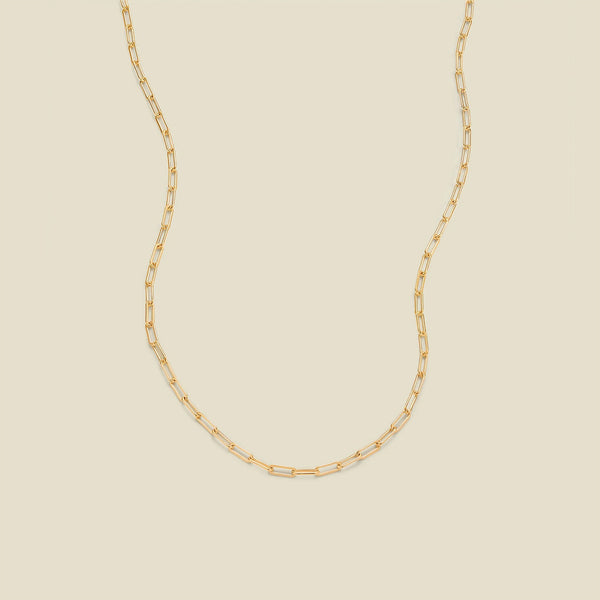 A gold paperclip chain necklace draped in a U-shape against a solid beige background.