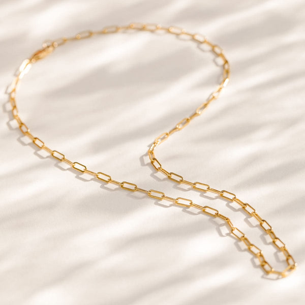 A gold paperclip link chain necklace laying on a textured white surface.