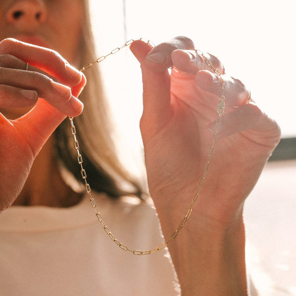 A person holding a gold paperclip link chain necklace in their hands, with sunlight coming through from behind them.