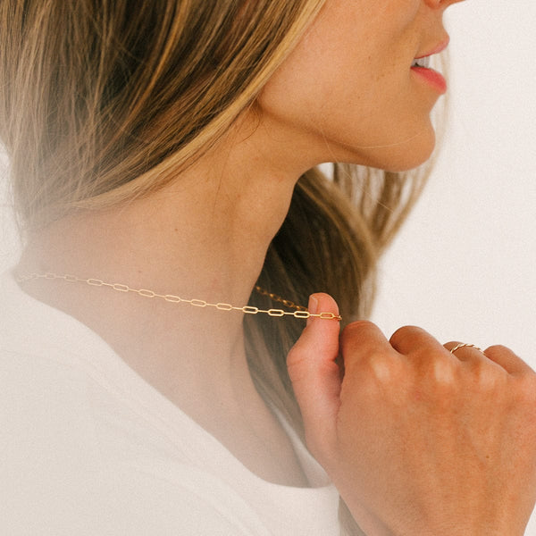 A person in a white top holding a gold paperclip link chain necklace out from their neck.
