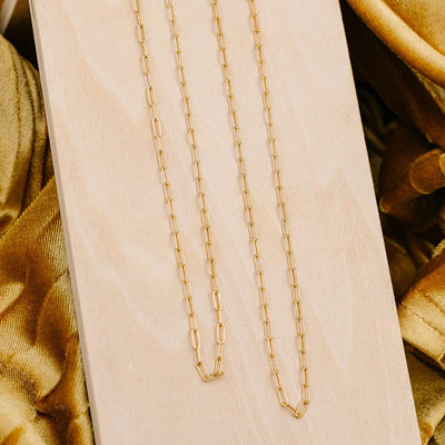 Two gold paperclip link chain necklaces in different lengths, draped on a light colored wood plank on top of golden fabric.