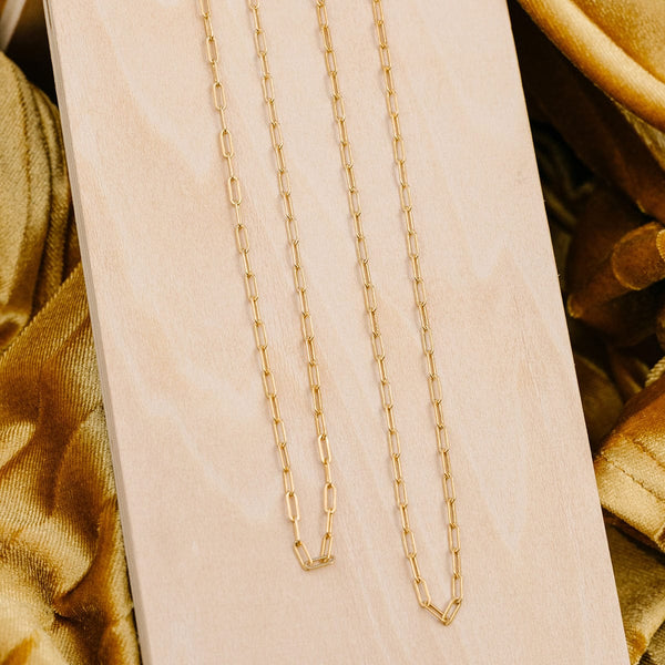 Two gold paperclip link chain necklaces in different lengths, draped on a light colored wood plank on top of golden fabric.