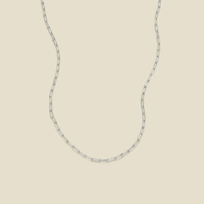 A silver paperclip chain necklace draped in a U-shape against a solid beige background.