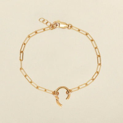 A gold paperclip link chain bracelet with a round ring in the center. The ring has a latch open, showing where to add charms to the bracelet, and it is shown against a solid light beige background.