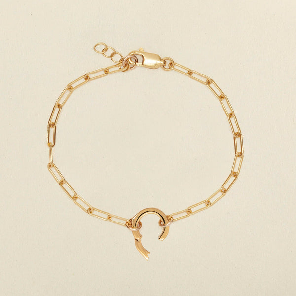 A gold paperclip link chain bracelet with a round ring in the center. The ring has a latch open, showing where to add charms to the bracelet, and it is shown against a solid light beige background.