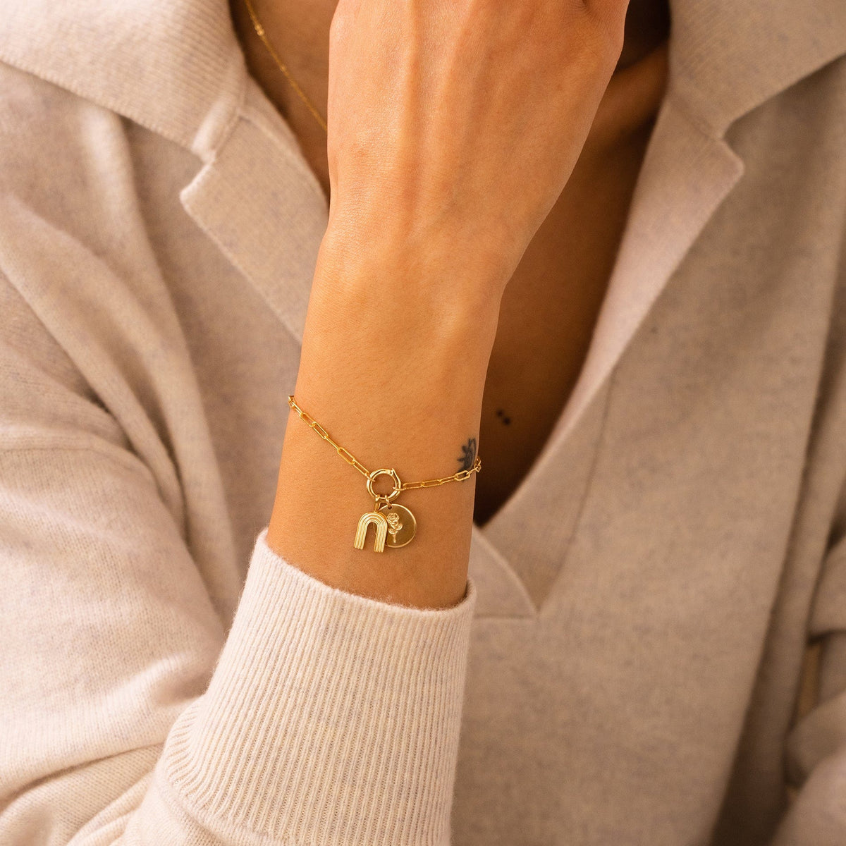 A person's wrist wearing a gold charm bracelet with a rainbow charm and flat round disc charm with a flower engraving.