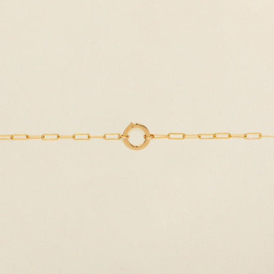 A gold paperclip link chain bracelet with a round ring in the center, shown against a solid light beige background.