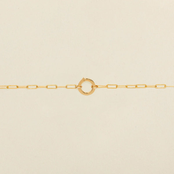 A gold paperclip link chain bracelet with a round ring in the center, shown against a solid light beige background.
