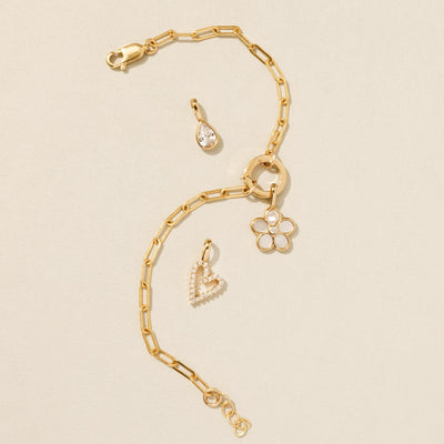 A gold paperclip link bracelet with a round ring in the center, holding a gold daisy charm with white inlay on the petals and a clear accent stone in the center. Laying around the bracelet are a pear shaped clear gemstone charm and a gold heart charm embellished with clear stones.