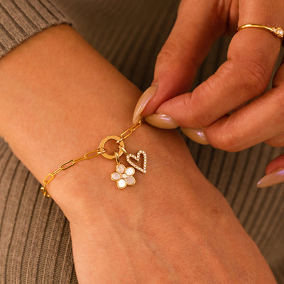 A close-up of a person's wrist wearing a gold paperclip link bracelet with a round ring in the center, holding a daisy charm with white inlay on the petals and a central clear accent stone and a gold heart charm embellished with clear stones.