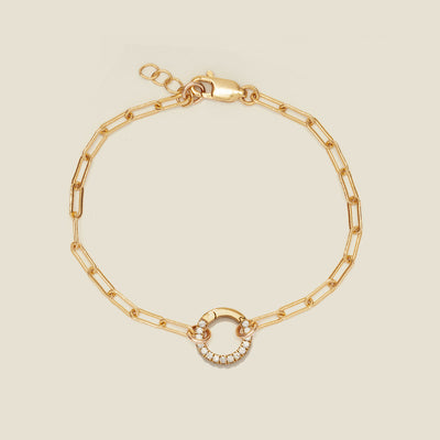 A gold paperclip link chain bracelet with a round ring in the center embellished with clear accent stones, shown on a solid light beige background.