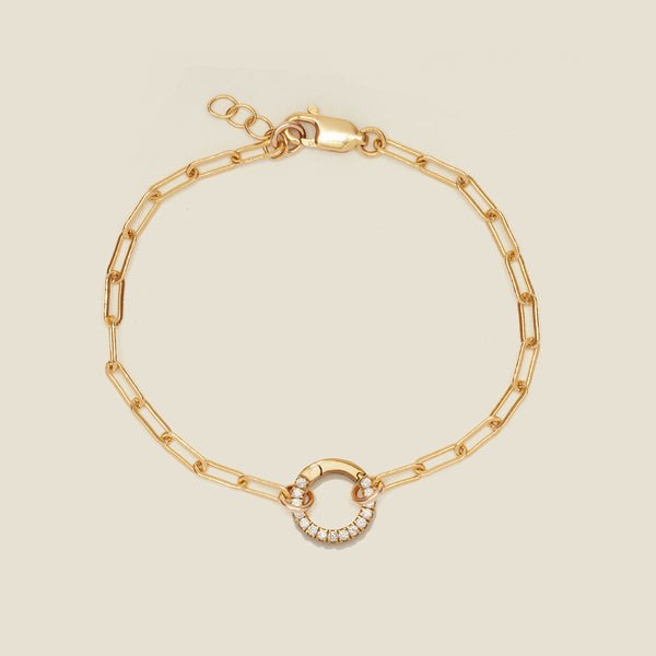 A gold paperclip link chain bracelet with a round ring in the center embellished with clear accent stones, shown on a solid light beige background.