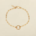A gold paperclip chain bracelet with a small circular ring in the center, displayed on a light beige background.