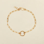 A gold paperclip chain bracelet with a small circular ring in the center, displayed on a light beige background.