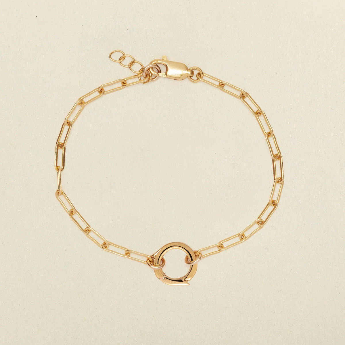 A gold paperclip chain bracelet with a small circular ring in the center, displayed on a light beige background.