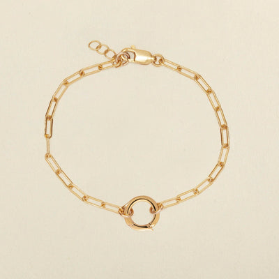 A gold paperclip chain bracelet with a small circular ring in the center, displayed on a light beige background.