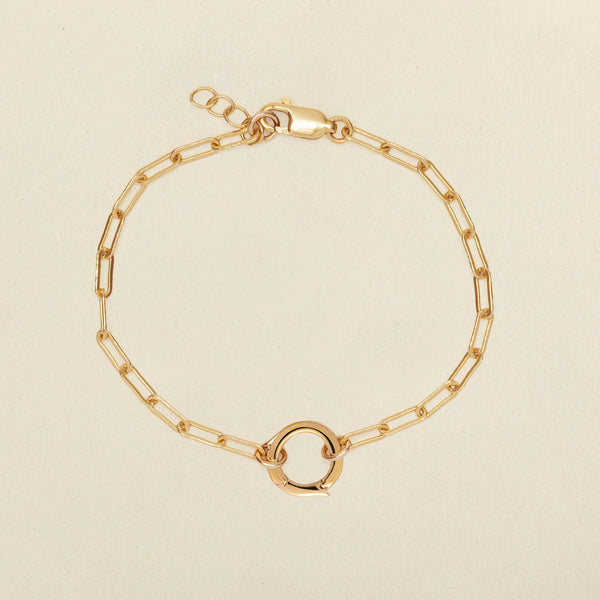 A gold paperclip chain bracelet with a small circular ring in the center, displayed on a light beige background.