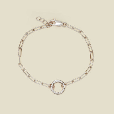 A silver paperclip link chain bracelet with a round ring in the center embellished with clear gemstones, shown against a solid light beige background.