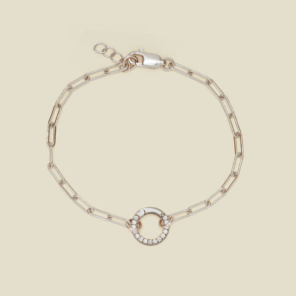 A silver paperclip link chain bracelet with a round ring in the center embellished with clear gemstones, shown against a solid light beige background.