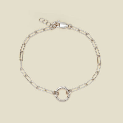 A silver paperclip link chain bracelet with a circular ring in the center, shown on a solid light beige background.
