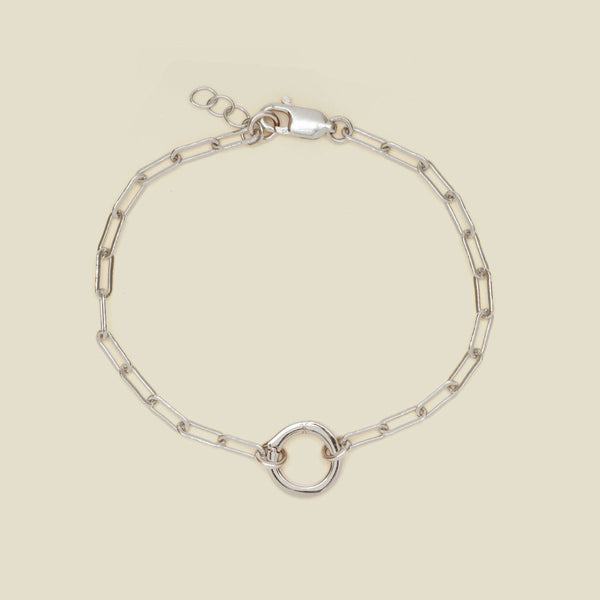 A silver paperclip link chain bracelet with a circular ring in the center, shown on a solid light beige background.
