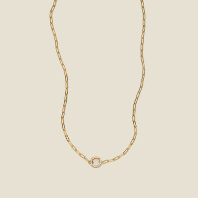 A gold paperclip link chain necklace with a round ring in the center embellished with clear gemstones, shown on a solid light beige background.
