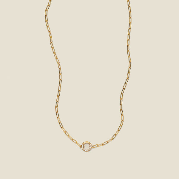 A gold paperclip link chain necklace with a round ring in the center embellished with clear gemstones, shown on a solid light beige background.