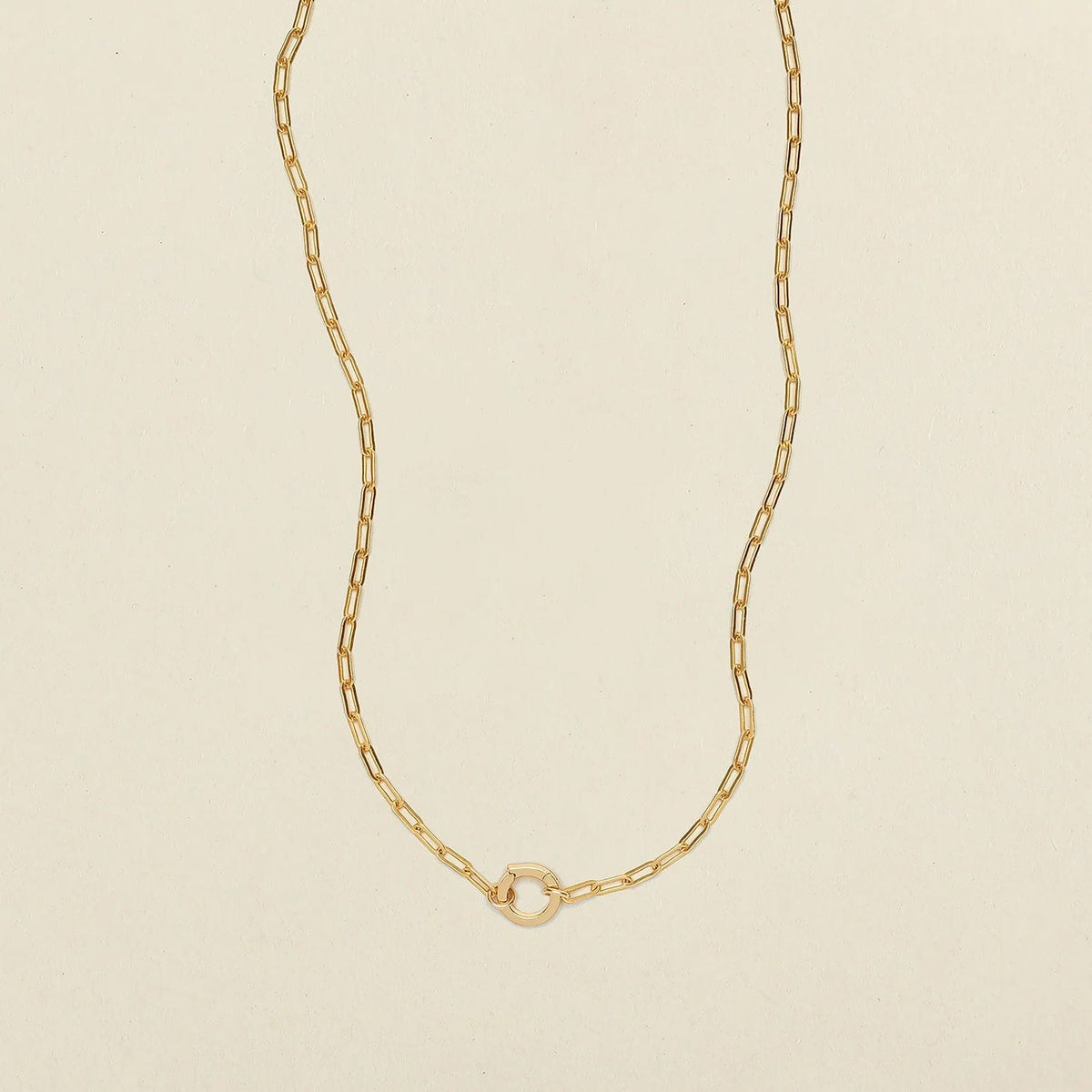 A delicate gold paperclip chain necklace with a small, polished gold ring in the center, displayed on a clean, light beige textured background.