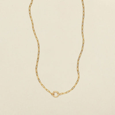 A delicate gold paperclip chain necklace with a small, polished gold ring in the center, displayed on a clean, light beige textured background.