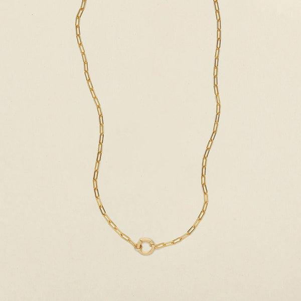 A delicate gold paperclip chain necklace with a small, polished gold ring in the center, displayed on a clean, light beige textured background.