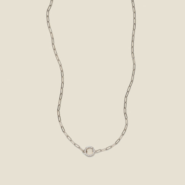 A silver paperclip link chain necklace with a round ring in the center embellished with clear accent stones, shown against a solid light beige background.