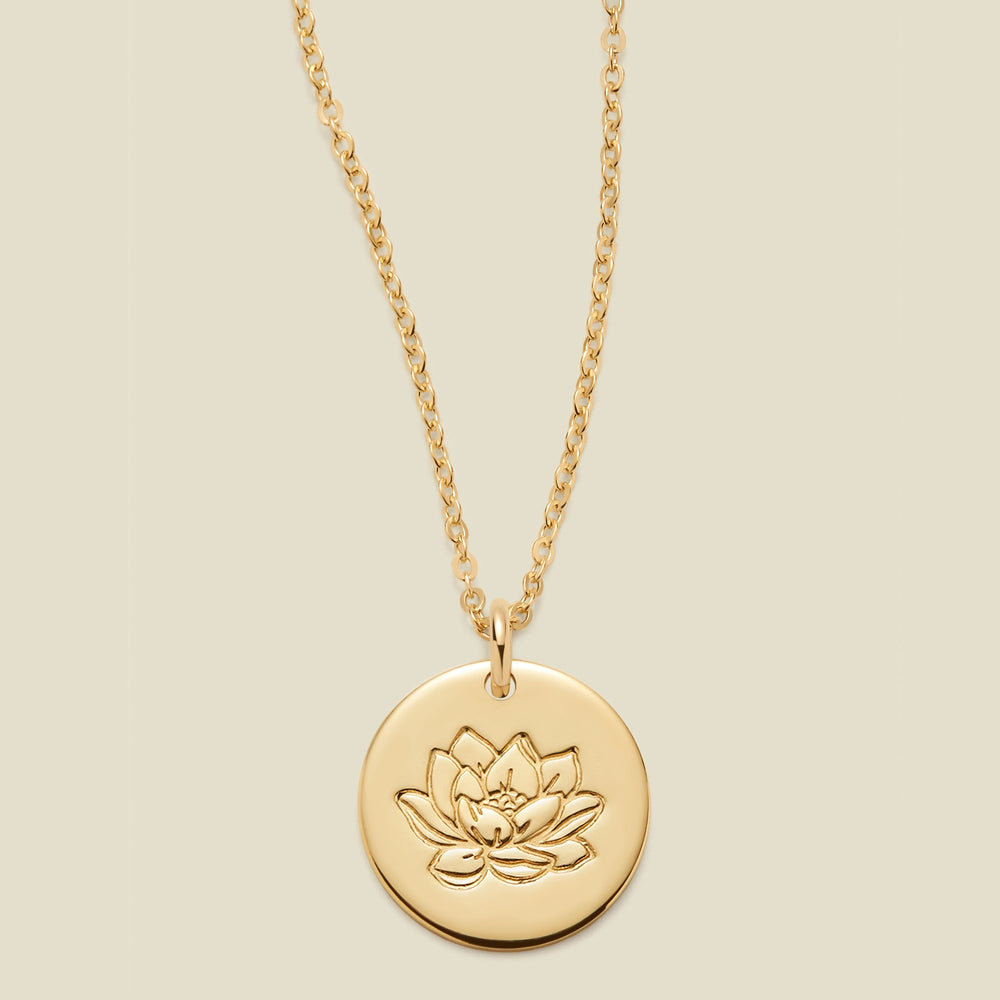 A gold chain necklace with a round gold disc pendant featuring an engraved lotus flower design, displayed against a solid light beige background.