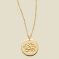 A gold chain necklace with a round gold disc pendant featuring an engraved lotus flower design, displayed against a solid light beige background.