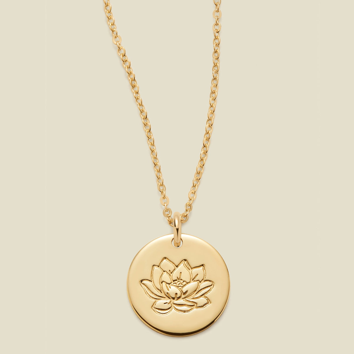 A gold chain necklace with a round gold disc pendant featuring an engraved lotus flower design, displayed against a solid light beige background.