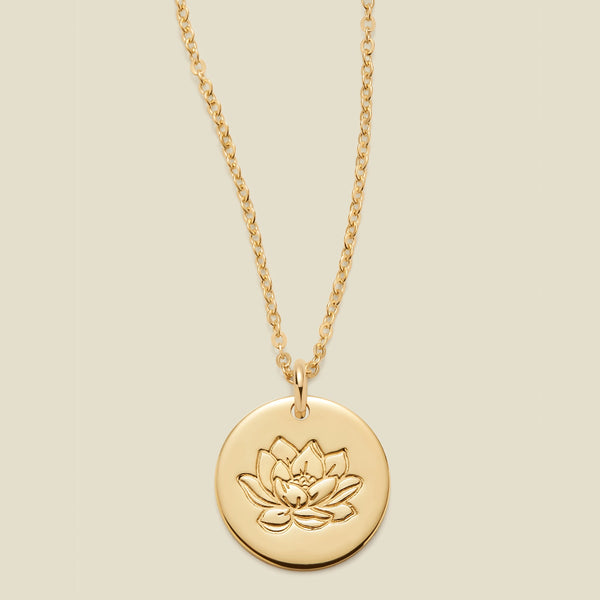 A gold chain necklace with a round gold disc pendant featuring an engraved lotus flower design, displayed against a solid light beige background.