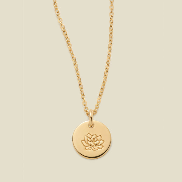 A gold chain necklace with a round gold disc pendant featuring an engraved lotus flower design, displayed against a solid light beige background.
