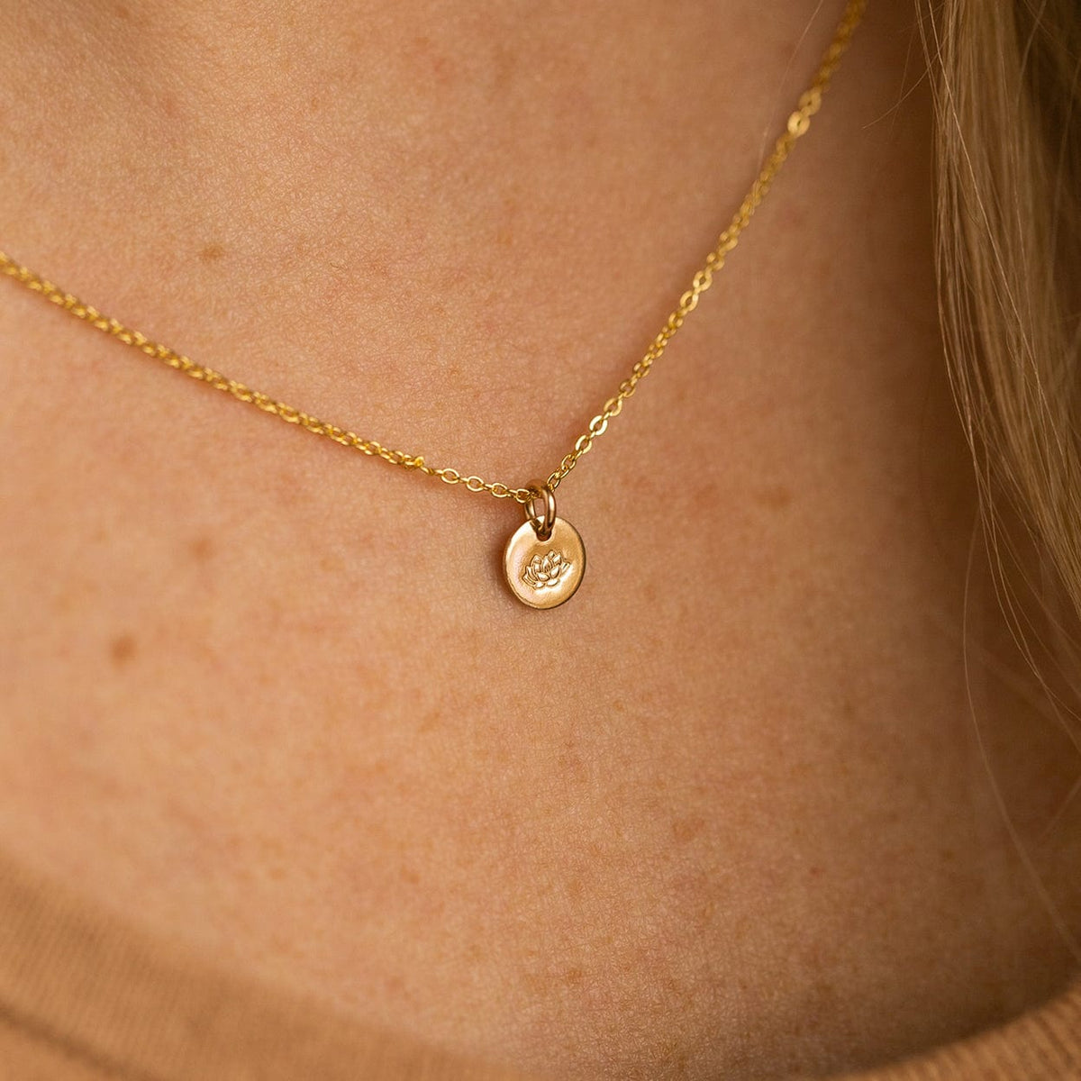 A close-up of a person wearing a delicate gold pendant necklace featuring a flat round disc charm, engraved with a lotus flower.