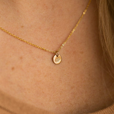 A close-up of a person wearing a delicate gold pendant necklace featuring a flat round disc charm, engraved with a lotus flower.