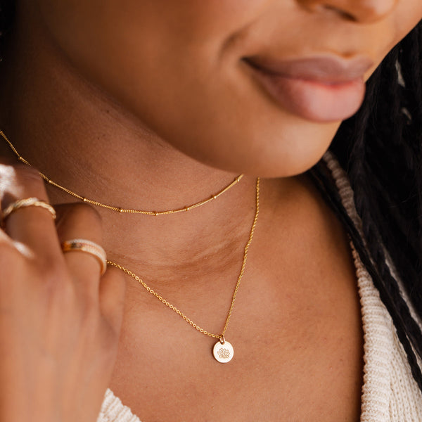 A close-up of a person wearing a thin gold choker necklace with stationary beads paired with a delicate gold pendant chain necklace featuring a round flat disc charm. The disc is engraved with a lotus flower design.