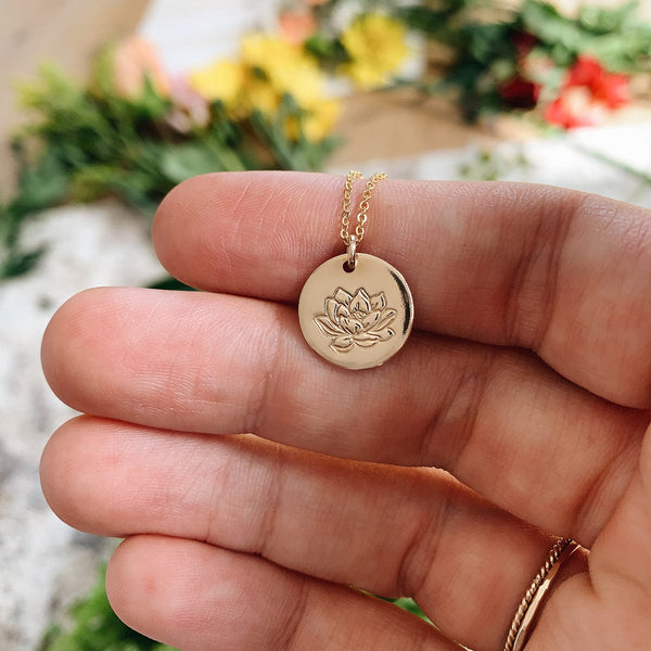 A hand holding a gold pendant chain necklace with a round flat disc charm, engraved with a lotus flower. The background is blurred with yellow, pink, and red flowers.