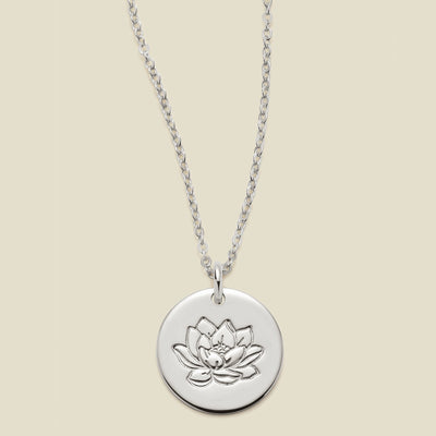 A silver chain necklace with a round silver disc pendant featuring an engraved lotus flower design, displayed against a solid light beige background.