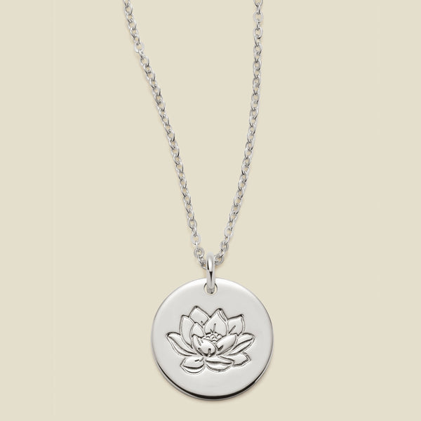 A silver chain necklace with a round silver disc pendant featuring an engraved lotus flower design, displayed against a solid light beige background.