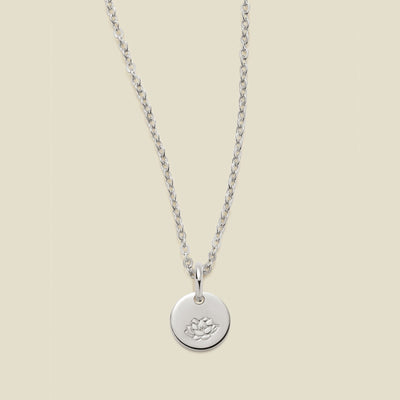 A silver chain necklace with a round silver disc pendant featuring an engraved lotus flower design, displayed against a solid light beige background.