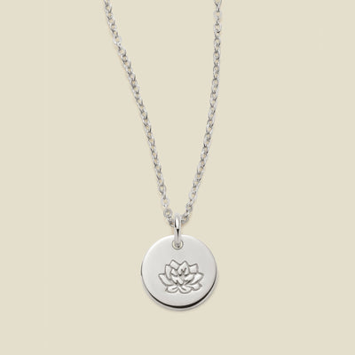 A silver chain necklace with a round silver disc pendant featuring an engraved lotus flower design, displayed against a solid light beige background.