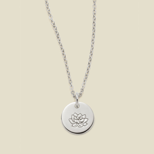 A silver chain necklace with a round silver disc pendant featuring an engraved lotus flower design, displayed against a solid light beige background.